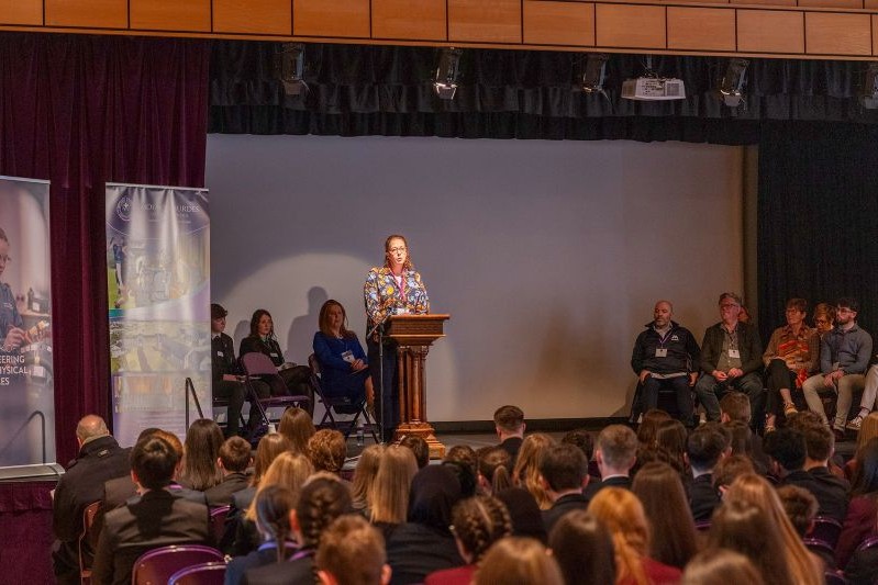 a woman with long red curly hair tied back stands at a lectern on a stage speaking to an audience of pupils
